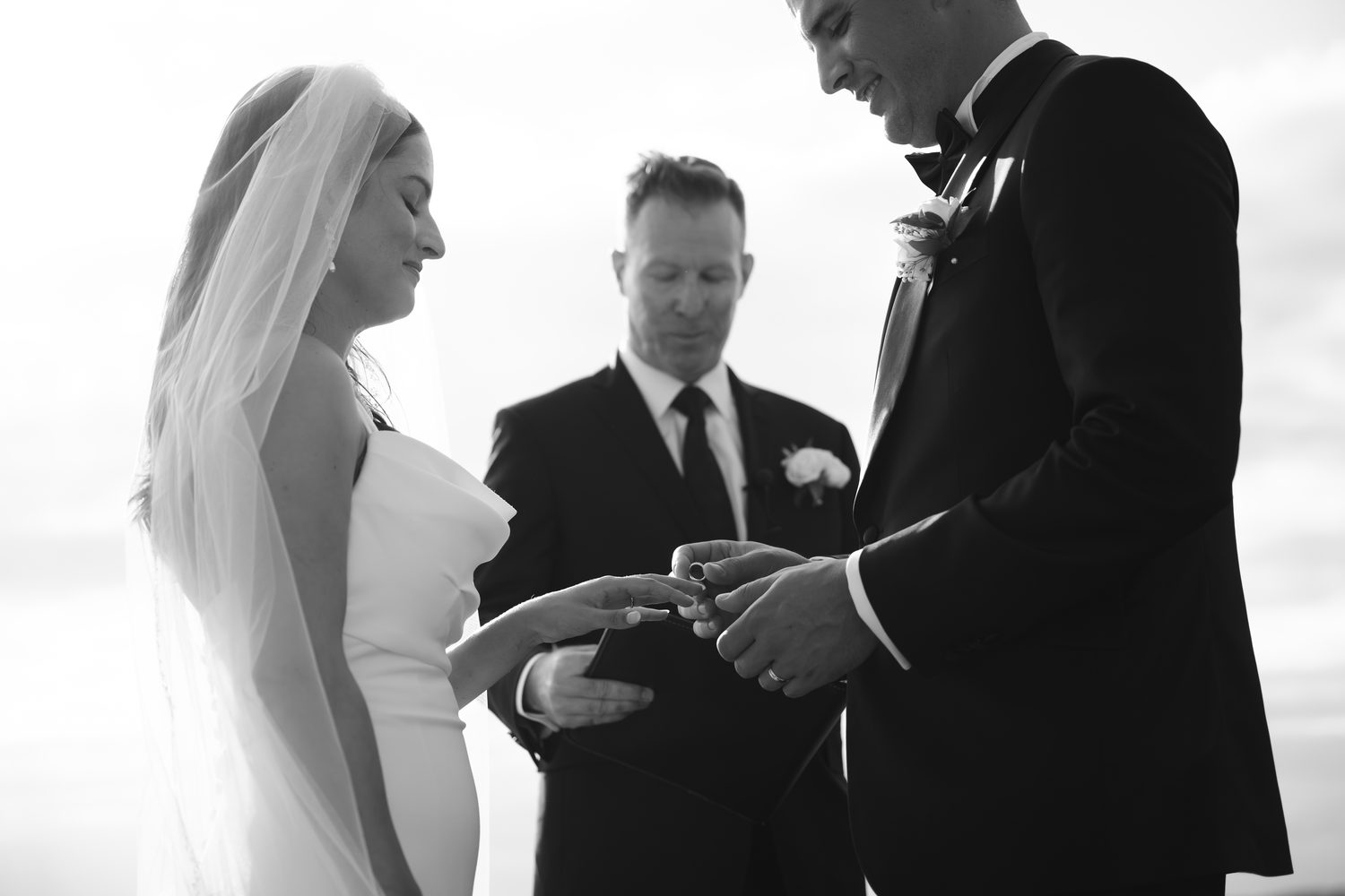 A couple exchanging rings during their ceremony as the officiant looks on, black and white